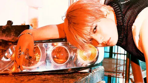 A person with bright orange hair leans over a car headlight in a warm, heavily filtered orange-toned setting.