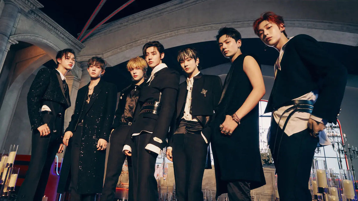 Seven male members of a music group pose together in stylish black outfits with white shirt details, set against a dark architectural background with warm lighting.