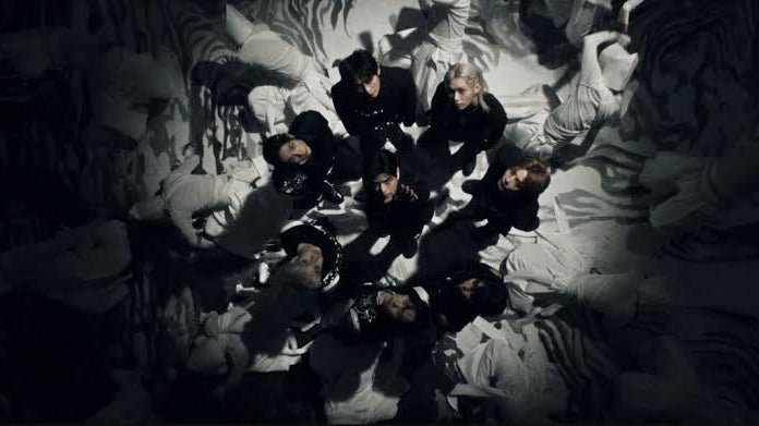 A group of people in black clothing gathered together in a dimly lit, monochrome scene with dramatic shadows and a textured, abstract background.