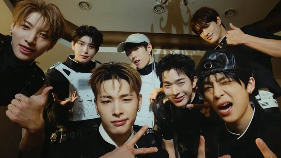 A group of seven young men posing closely together indoors, wearing mostly black outfits with some white and gray accents. The setting appears to be a dimly lit room with a chandelier visible overhead.