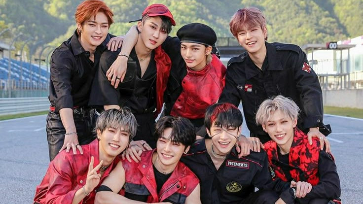 Eight male members of a K-pop group pose together on a racetrack, wearing coordinated black and red outfits. The background shows green hills and the racetrack surface.