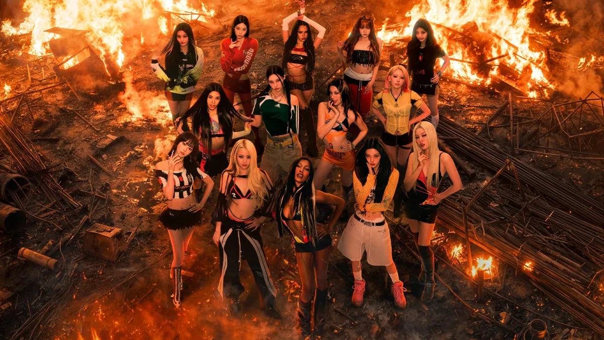 Fourteen women posing together in a fiery, debris-filled outdoor scene. They are dressed in varied streetwear and fashion styles, with bright orange flames and smoke filling the background.