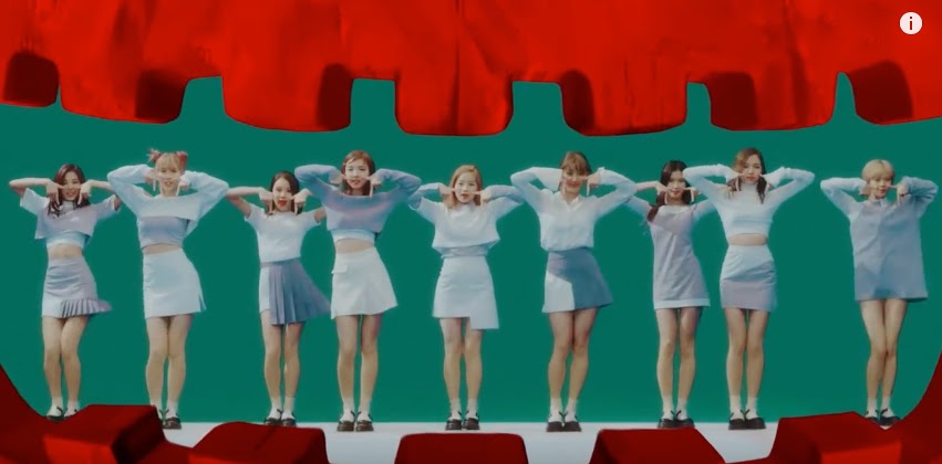 Nine women in matching light blue and white outfits pose together in front of a green screen with a red curtain backdrop.
