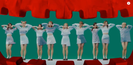 Nine women in matching light blue and white outfits pose together in front of a green screen with a red curtain backdrop.