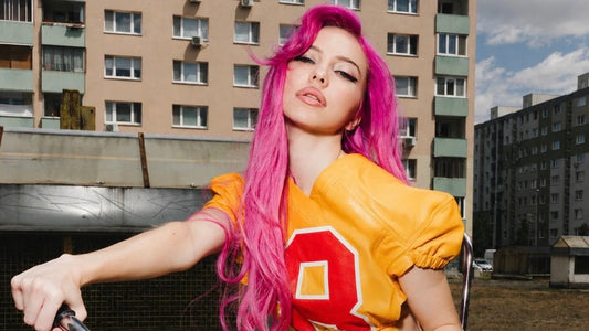 A woman with vibrant pink hair poses outdoors on a metal structure, wearing a yellow short-sleeve football-style top with a large red number 8 on the front, paired with a white skirt.