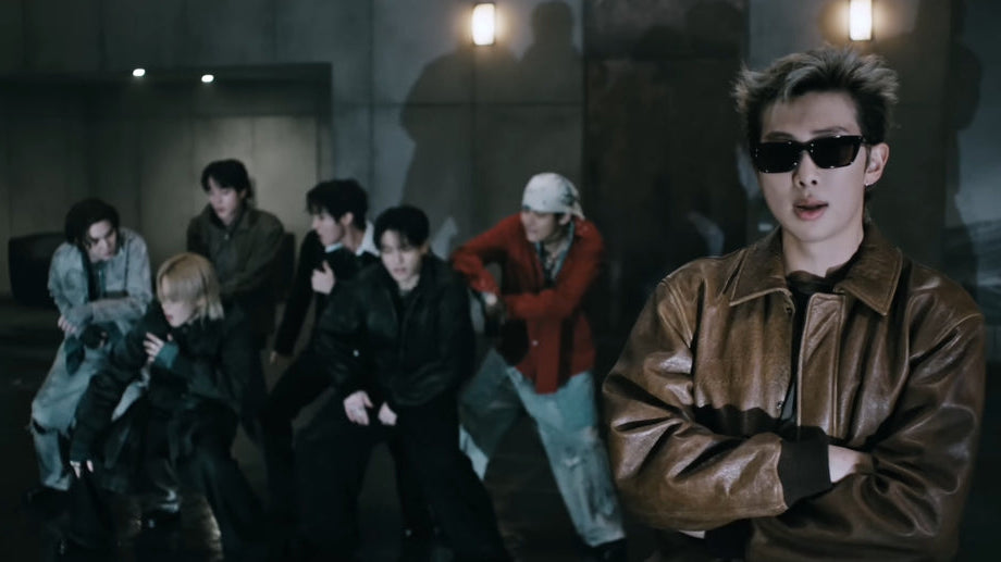 A man in a brown leather jacket and black sunglasses stands in the foreground with arms crossed, while a group of people in dark clothing poses in a dimly lit indoor setting behind him.