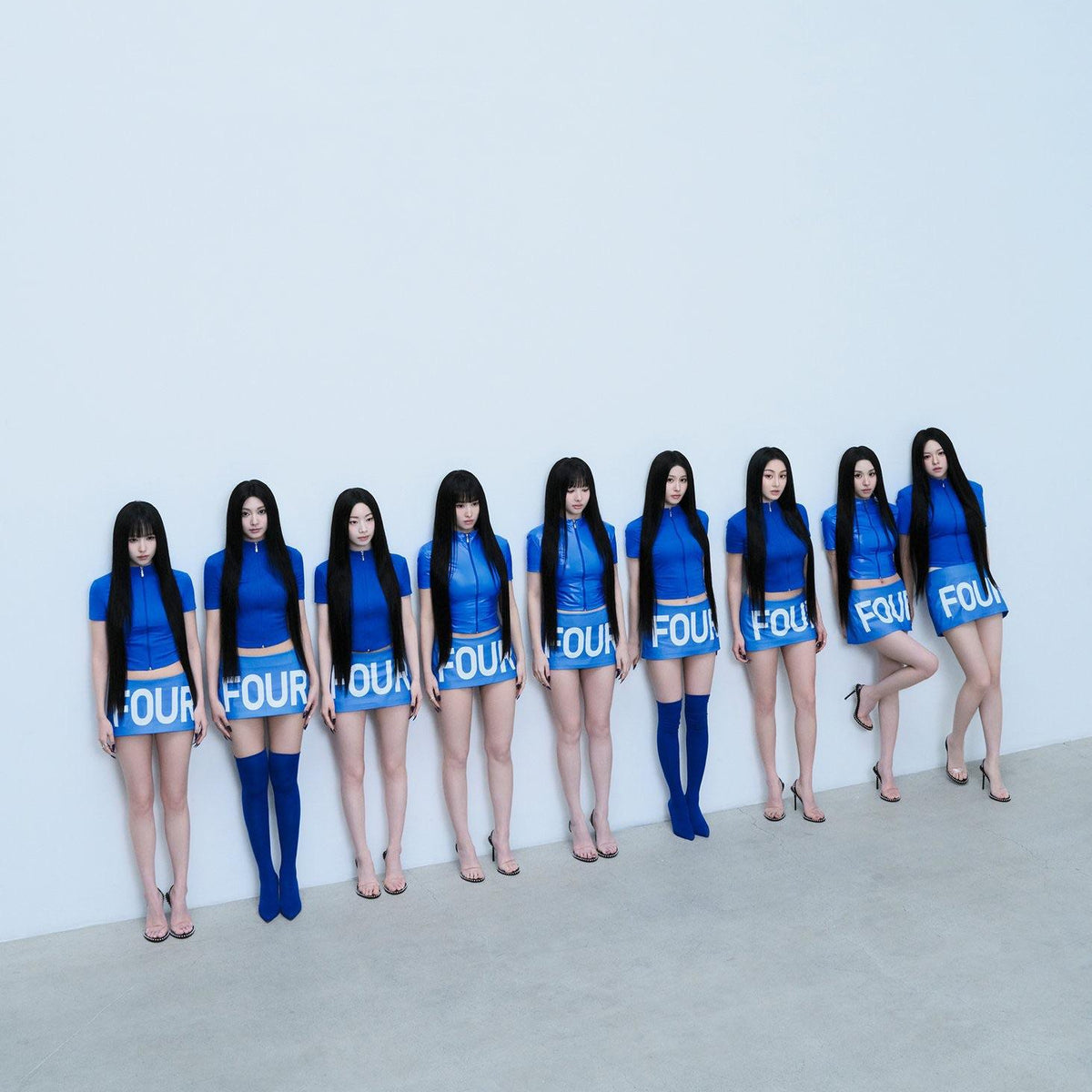 Nine women stand in a row against a plain white wall wearing matching outfits consisting of a blue short-sleeve top with 'FOUR' printed in white, a matching blue mini skirt, and either blue thigh-high socks or nude heels.