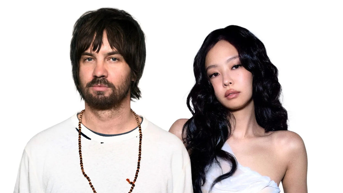 A man with dark hair and a beard wearing a white t-shirt and a beaded necklace, standing next to a woman with long black hair wearing a white strapless top, against a solid white background.